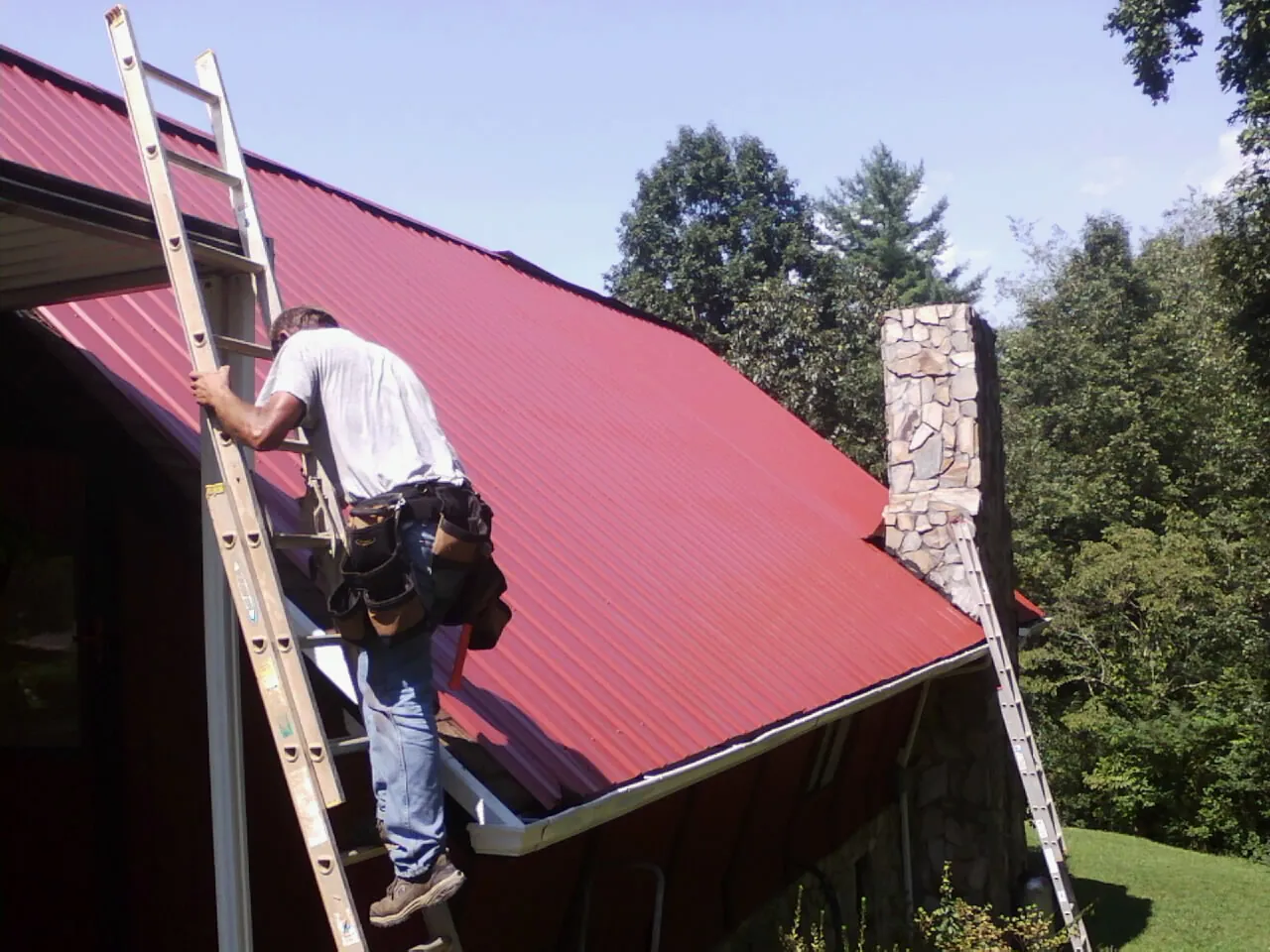Roofer on a ladder beside a red metal roof and stone chimney — Soffit & Fascia Repair by Roofing Brothers in Shaw Afb